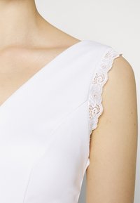 Vestido branco com decote em V e delicadas rendas nos ombros, feito de um tecido suave com um silhueta elegante.