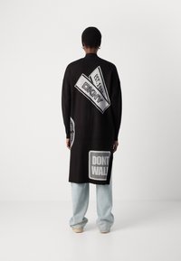 Duga crna jakna s grafičkim aplikacijama, uključujući "DKNY" i "NE HODAJ", u kontrastnoj bijeloj boji. Širokog kroja, s visokim bočnim prorezima i širokim rukavima.