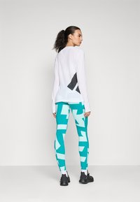 Haut à manches longues blanc avec dos en mesh et accents noirs, associé à un legging teal avec des motifs géométriques et des chaussures noires.