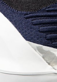 Obuv s tmavě modrým pleteným svrškem s texturovanými akcenty, přecházející do hladké bílé gumové podrážky. Viditelné jemné detaily camo vzoru.