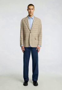 Beež kootud bleiser ühe nupu kinnihoidmisega, ühendatud helesinise särk ja tumedate siniste pükstega. Mudel seisab neutraalses taustas.