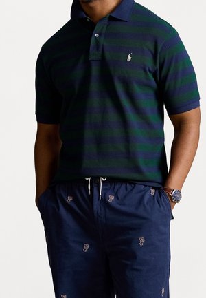 Personne portant un polo à rayures vertes et bleu marine avec un logo brodé, un short bleu marine avec de petits logos à motifs, et une montre-bracelet en cuir marron.