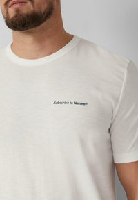Witte, kortgemouwde t-shirt van zachte stof met een ronde hals. Bevat groene tekst "Abonneer op de Natuur®" op de linkerborst.