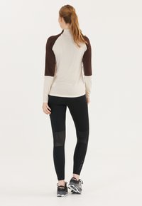 Langærmet bluse i lys beige med mørkebrune raglanærmer; parret med sorte leggings med mesh-detaljer og ankel-detaljer.