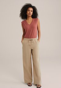 Top fără mâneci, cu decolteu în V, de culoare roz, combinat cu pantaloni largi bej. Pantalonii au talie elastică și șnur de ajustare, cu o textură de țesătură netedă.