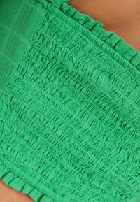 Zielona bluzka z tkaniny o marszczonym wykończeniu z efektem pogniecenia oraz falbaniastym dekoltem. Materiał wydaje się lekki i przepuszczający powietrze.