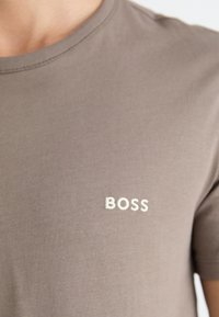 T-shirt en coton marron avec un col rond, featuring un logo "BOSS" brodé en blanc sur la partie gauche de la poitrine. Texture lisse.