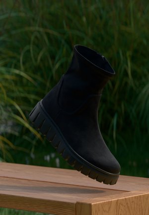 Bottine noire à semelle épaisse rainurée flottant au-dessus d'une table en bois, fond d'herbe verte.