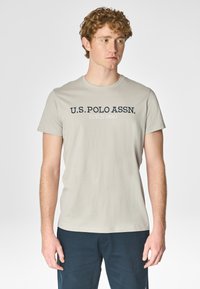 T-shirt grigio chiaro con collo a giro, con la scritta "U.S. POLO ASSN." stampata in blu navy sul petto. Materiale in morbido cotone misto.