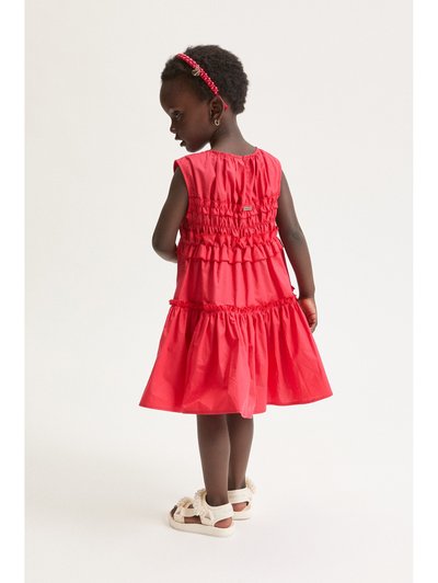 Jeune fille aux cheveux courts et bouclés portant une robe rouge à volants, un bandeau rouge, des boucles d'oreilles créoles dorées et des sandales blanches ornées de perles décoratives.