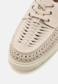 Zapato de ante beige con textura trenzada, que presenta un detalle trenzado en el borde y ojales redondos para los cordones, con una suela de goma blanca.