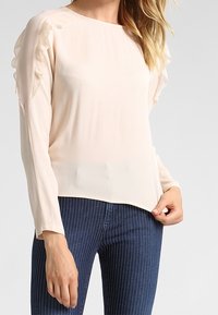 Blusa rosa chiaro con maniche lunghe, caratterizzata da dettagli arricciati sulle spalle e una scollatura rotonda, abbinata a jeans a strisce scure.