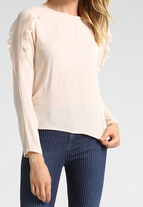 Blusa rosa chiaro con maniche lunghe, caratterizzata da dettagli arricciati sulle spalle e una scollatura rotonda, abbinata a jeans a strisce scure.