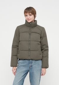Kvinde med kort hår iført grøn polstret jakke og lyseblå jeans, stående foran en ensfarvet hvid baggrund.