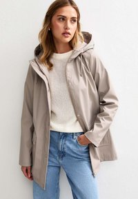 Veste imperméable beige avec capuche, dotée d'une fermeture éclair à l'avant, de poches latérales et d'une coupe décontractée, superposée à un pull en tricot crème et un jean bleu.