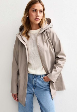 Veste imperméable beige avec capuche, dotée d'une fermeture éclair à l'avant, de poches latérales et d'une coupe décontractée, superposée à un pull en tricot crème et un jean bleu.