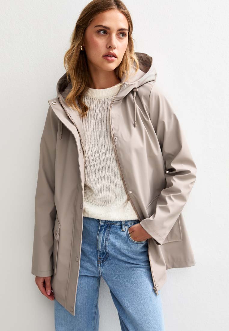 Veste imperméable beige avec capuche, dotée d'une fermeture éclair à l'avant, de poches latérales et d'une coupe décontractée, superposée à un pull en tricot crème et un jean bleu.