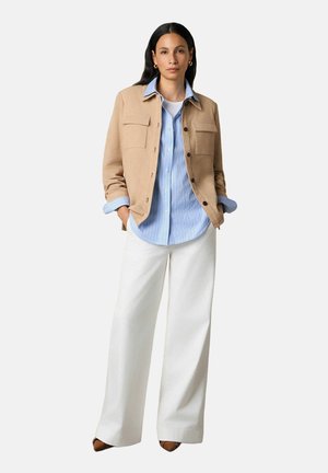 Frau steht mit den Händen in den Taschen, trägt eine beige Jacke, ein blau gestreiftes Hemd, weiße Hosen mit weitem Bein und braune spitze Schuhe.