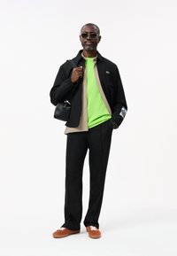 Zwarte jack over een groen T-shirt, gestreept shirt eronder, zwarte broek, bruine sneakers met witte veters, zwarte tas, zonnebril.