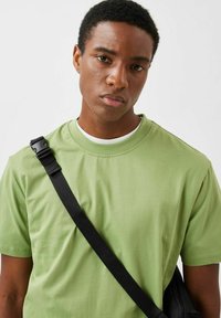 Grøn bomulds T-shirt med rund hals, kortærmede og afslappet pasform, båret med en sort skuldertaske.