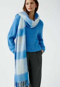 Pull en tricot bleu à coupe décontractée, associé à une écharpe épaisse avec un motif à carreaux dégradés bleu et blanc et des franges aux extrémités.