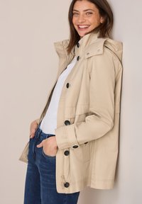 Vedenpitävä beige takki hupulla, jossa on mustat napit, taskut läpällä ja rento istuvuus. Päällä valkoinen toppi ja siniset farkut.