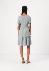Sundress azul claro com nervuras, mangas curtas, bainha em camadas e detalhes com bordas desfalcadas. Combinado com sapatos de salto bejes com tiras. Manequim visto de trás.