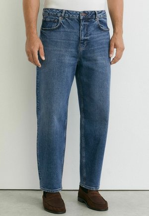 Homme portant un jean bleu droit avec une braguette à boutons et fermeture éclair, associé à des chaussures marron sans lacets sur un sol clair.
