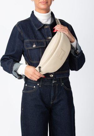 Beige læder skuldertaske med en rundet form, der har lynlås, logo-patch og en justerbar rem, holdt op mod en mørk denimoutfit.