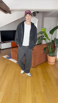 Μαύρη ζακέτα με φερμουάρ, λευκό μπλουζάκι, γκρι φόρμα και ασημένια αθλητικά παπούτσια. Εσωτερικός χώρος με καφέ καναπέ και φυτά σε γλάστρες. Ξύλινο πάτωμα.