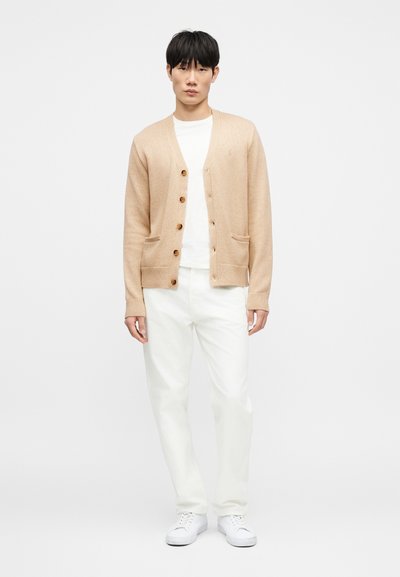 Μπεζ ζακέτα με V-λαιμόκοψη, πέντε κουμπιά και δύο μπροστινές τσέπες. Φορεμένη πάνω από ένα λευκό μπλουζάκι και συνδυασμένη με λευκό παντελόνι και αθλητικά παπούτσια.