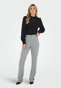 Femme debout avec des cheveux blonds ondulés, portant un haut en dentelle noire à manches longues, un pantalon gris taille haute et des chaussures pointues noires.
