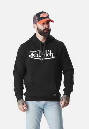 Homme barbu portant un sweat à capuche noir Von Dutch, une casquette orange et noire Von Dutch, un jean bleu clair, et montrant ses mains tatouées.
