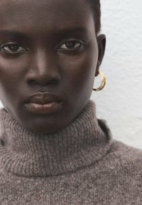 Šedý žebrovaný rolák s vysokým límcem. Zlaté kruhové náušnice. Hladká pleť s přirozeným líčením, neutrální pozadí.