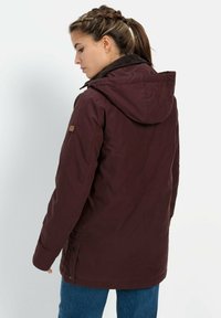 Bordeauxjacke mit Kapuze, aus weichem Material, mit Knöpfen vorne und einem seitlichen Logo-Patch, getragen über blauen Jeans.