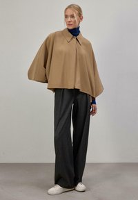Veste cape kaki avec un col montant, fermeture par boutons et manches amples, associée à un pantalon à carreaux gris foncé et des baskets blanches.