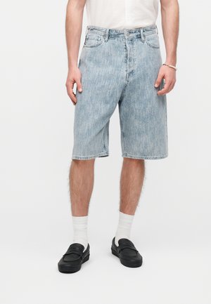 Man wearing light blue denim knee-length shorts, white socks, black slip-on shoes, and a white t-shirt, standing against a plain background.