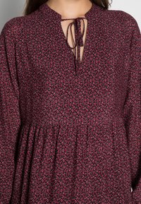 Långärmad klänning i mörkröd tyg med blomsterdesign, med en nyckelhålsklänningskrage som är knuten med en rosett och en sammangjord midja.