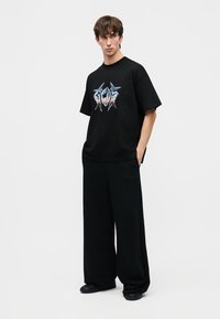 Νέος άνδρας φορώντας μαύρο φαρδύ παντελόνι και μαύρο T-shirt με γραφικά, στέκεται με τα χέρια στις τσέπες, κοιτάζοντας προς τα πλάγια.