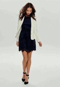 Jauna moteris, pasitikinti savimi, vilkinti tamsiai mėlyną nėriniuotą suknelę, baltą švarką ir juodas aukštakulnes batais su kulkšną raiščiu, stovinti ant šviesaus vientiso fono.