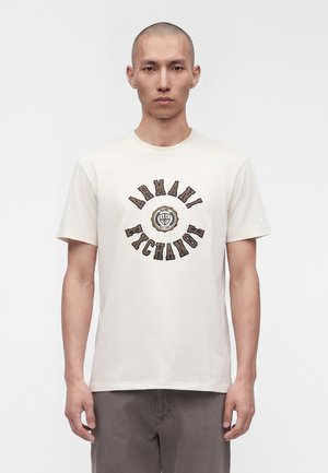 Νέος άνδρας που φοράει λευκό μπλουζάκι Armani Exchange με καφέ λογότυπο και έμβλημα, συνδυασμένο με γκρι παντελόνι, στέκεται μπροστά σε απλό λευκό φόντο.