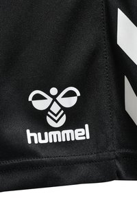 Le tissu du maillot de sport noir présente un logo Hummel blanc avec un design d'abeille et des rayures blanches diagonales sur le côté. Texture lisse.