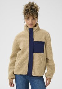 Veste en polaire beige avec un col montant, dotée d'une poche poitrine zippée bleu marine et d'un tissu doux et texturé. Coupe décontractée avec des manches longues.