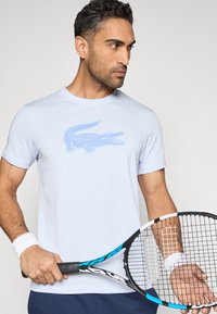 Gaiši zils kokvilnas T-krekls ar lielu zilu krokodila logotipu uz krūtīm, kombinēts ar tenisa raketi vīrieša sportiskā pozā.