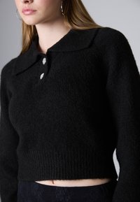 Pulover negru din tricot cu guler, având trei butoane decorative și un design scurt. Textură moale și detaliu cu margine ondulată.