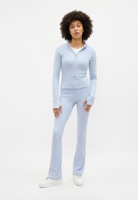 Tuta di colore blu chiaro realizzato in un tessuto liscio e elasticizzato. Presenta una giacca con zip e pantaloni a campana, entrambi con maniche lunghe e polsini.