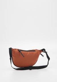 Brązowa nylonowa torba na ramię o wygiętym kształcie, z regulowanym czarnym paskiem i zapinaną na zamek kieszenią z przodu. Gładka tekstura.