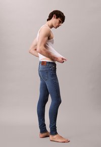 Fiatal férfi mezítláb, kék szűk farmernadrágot és fehér ujjatlan pólót visel, amelyet elhúz a testétől.