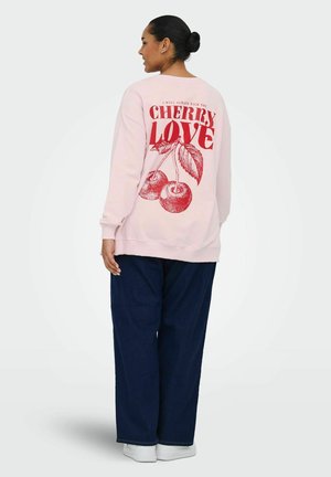 Femme aux cheveux attachés en chignon, portant un sweat-shirt rose clair avec le texte rouge "Cherry Love" et une illustration de cerise, un pantalon bleu foncé et des chaussures blanches.