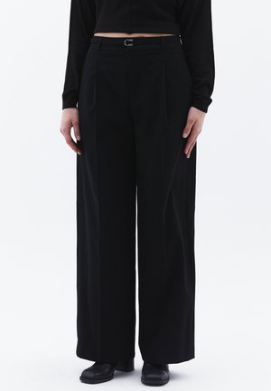 Pantalons larges noirs en tissu lisse, avec des plis devant, une taille ajustée et un petit détail de boucle en argent.